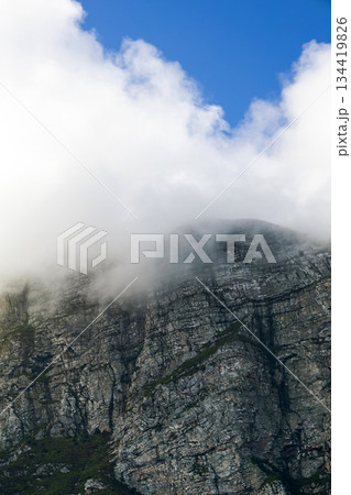 Majestic mountain peak emerging from a layer of clouds, rugged rock formation 134419826