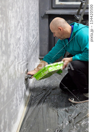 A focused bald man is carefully applying decorative plaster to a wall in a room 134420500