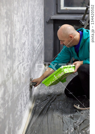 Close-up of a focused man applying textured plaster to a wall with a green tray and trowel 134420502