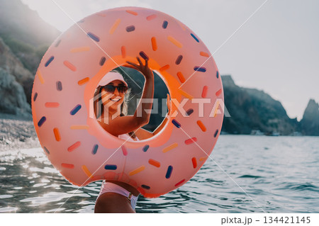 Woman, donut, sea. Happy woman holds pink donut inflatable ring in sparkling sea during summer vacation with copy space. Woman, donut, sea. Happy woman holds pink donut inflatable ring in sparkling sea during summer vacation with copy space. 134421145