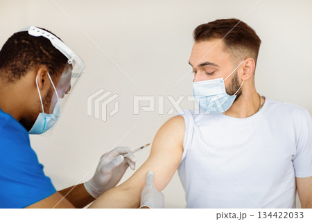 Young Caucasian man getting injection of coronavirus vaccine at hospital. African American medical worker immunizing male patient against covid-19 at clinic. Healthcare and global infection prevention Young Caucasian man getting injection of coronavirus vaccine at hospital. African American medical worker immunizing male patient against covid-19 at clinic. Healthcare and global infection prevention 134422033