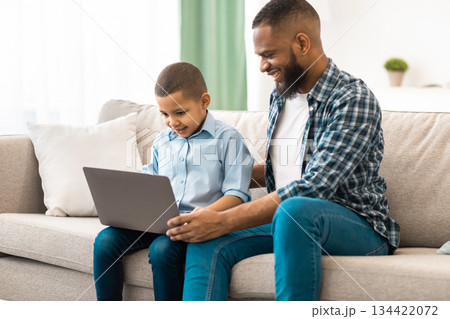 African Father And Son Using Laptop Browsing Internet Together Sitting On Couch At Home. Weekend With Dad. Little Boy And His Dad Watching Movie On Computer Online. Side View 134422072