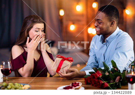 Romantic Surprise. Affectionate Black Man Giving Present To His Excited White Girlfriend, Greeting Her With International Women's Day, Happy Interracial Couple Celebrating On Date In Restaurant Romantic Surprise. Affectionate Black Man Giving Present To His Excited White Girlfriend, Greeting Her With International Women's Day, Happy Interracial Couple Celebrating On Date In Restaurant 134422089