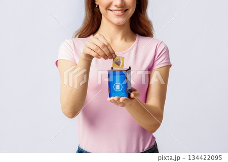 Trash Recycling. Young Woman Putting Card With Paper Waste Icon Into Blue Wooden Toy Garbage Container, Standing Over Light Studio Background, Conceptual Image For Recycle, Copy Space Trash Recycling. Young Woman Putting Card With Paper Waste Icon Into Blue Wooden Toy Garbage Container, Standing Over Light Studio Background, Conceptual Image For Recycle, Copy Space 134422095