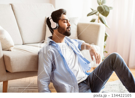 Time To Relax. Calm arab man in wireless headphones listening music at home, resting on floor in living room, sitting with closed eyes and enjoying favorite melodies, stress relief concept, free space Time To Relax. Calm arab man in wireless headphones listening music at home, resting on floor in living room, sitting with closed eyes and enjoying favorite melodies, stress relief concept, free space 134422105