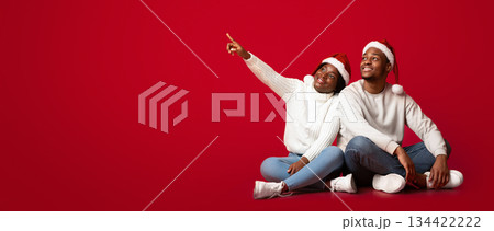 Holiday Offer. Interested Black Man And Woman In Santa Hats Looking And Pointing Away At Copy Space On Orange Background, Sitting African Couple Excited About Christmas Sale Or Promo, Panorama Holiday Offer. Interested Black Man And Woman In Santa Hats Looking And Pointing Away At Copy Space On Orange Background, Sitting African Couple Excited About Christmas Sale Or Promo, Panorama 134422222