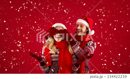 Affectionate young man in Christmas outfit closing his girlfriend's eyes on red background Affectionate young man in Christmas outfit closing his girlfriend's eyes on red background 134422239