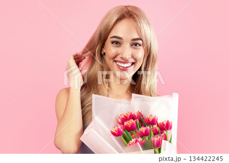 Gorgeous lady in lovely dress holding tulips, smiling and looking at camera over pink studio background. Beautiful blonde model posing with spring flowers. Special gift for Woman's Day Gorgeous lady in lovely dress holding tulips, smiling and looking at camera over pink studio background. Beautiful blonde model posing with spring flowers. Special gift for Woman's Day 134422245