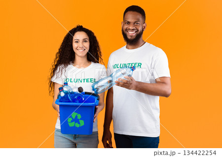 Two Cheerful Volunteers Holding Box With Recycling Symbol Full Of Wasted Plastic Bottles Standing On Yellow Background In Studio. Garbage Disposal, Sort And Recycle Your Waste Concept Two Cheerful Volunteers Holding Box With Recycling Symbol Full Of Wasted Plastic Bottles Standing On Yellow Background In Studio. Garbage Disposal, Sort And Recycle Your Waste Concept 134422254
