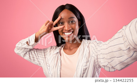 Carefree young black woman taking selfie and showing peace gesture on pink studio background. Positive African American lady smiling at camera and making photo of herself on mobile phone 134422255