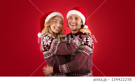 Expressing affection. Lovely young couple in Santa hats and Christmas sweaters hugging on red background 134422256
