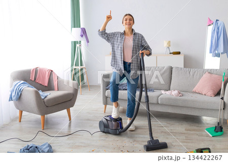 Happy young lady with vacuum cleaner having cleanup idea at messy room after party. Millennial female student hoovering dirty apartment, pointing finger up, gesturing eureka Happy young lady with vacuum cleaner having cleanup idea at messy room after party. Millennial female student hoovering dirty apartment, pointing finger up, gesturing eureka 134422267