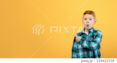 Amazed red-haired school boy in plaid shirt aiming at copy space over yellow studio background. Shocked ginger kid pointing at empty space, showing exciting advert for children 134422319