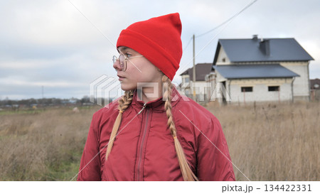 Young girl sporting braided hair, red beanie, winter jacket, gazing across rural landscape with unfinished residential construction site behind her 134422331