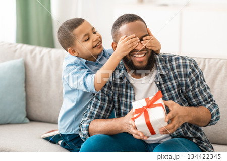Dad's Birthday. Little Son Congratulating Black Father Giving Wrapped Gift To Him, Covering Eyes Sitting On Couch At Home. Family Holiday Celebration, Surprise Present For Daddy's B-Day 134422345