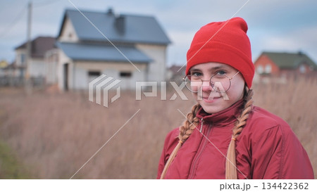 Teenage girl smiling, wearing red beanie and glasses, standing in grassy field with residential neighborhood behind Teenage girl smiling, wearing red beanie and glasses, standing in grassy field with residential neighborhood behind 134422362