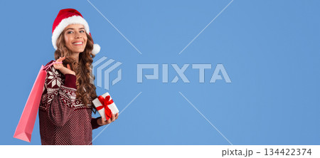 Pretty lady in winter outfit holding Christmas gift and shopping bag on blue background, panorama Pretty lady in winter outfit holding Christmas gift and shopping bag on blue background, panorama 134422374