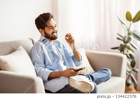 Watching TV At Home. Cheerful arab guy resting on couch and eating popcorn, smiling millennial eastern man enjoying his favorite television show, having domestic leisure, copy space 134422376