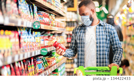 Grocery Shopping. African American Guy Buying Food In Supermarket Store Wearing Protective Face Mask, Standing With Cart In Shop. Male Buyer Customer Choosing Products. Copy Space 134422433