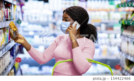 Lovely African American woman black lady in face mask calling on cellphone while shopping for food at modern supermarket. Young black lady buying groceries at mall during coronavirus pandemic Lovely African American woman black lady in face mask calling on cellphone while shopping for food at modern supermarket. Young black lady buying groceries at mall during coronavirus pandemic 134422466