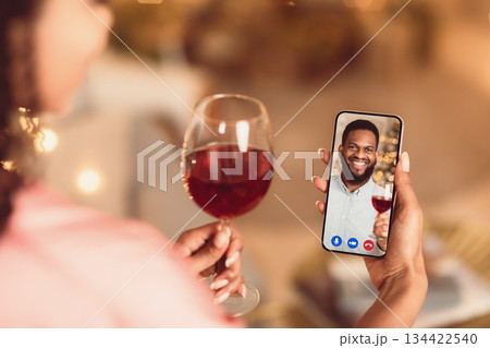 Celebrating Anniversary. Back over the shoulder view of young black woman having a romantic online date with her man, holding glass of red wine and mobile phone, toasting. Stay Safe At Home Celebrating Anniversary. Back over the shoulder view of young black woman having a romantic online date with her man, holding glass of red wine and mobile phone, toasting. Stay Safe At Home 134422540