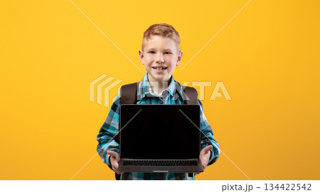 Cheerful ginger kid with schoolbag showing laptop with blank screen on yellow studio background, copy space. Happy schoolboy holding modern laptop, using technologies in education, mockup 134422542