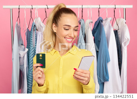 Online Payment Concept. Portrait of smiling stylish lady holding credit card and using mobile phone for shopping in internet, ordering clothes, standing with rack in the pink background Online Payment Concept. Portrait of smiling stylish lady holding credit card and using mobile phone for shopping in internet, ordering clothes, standing with rack in the pink background 134422551