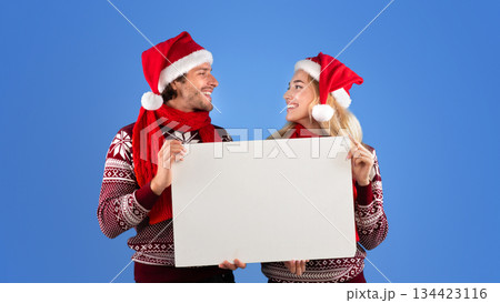 Happy young couple in Christmas outfits holding empty banner with mockup for design over blue background Happy young couple in Christmas outfits holding empty banner with mockup for design over blue background 134423116
