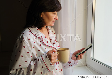 Woman in pajamas drinking coffee and using smartphone near window in morning light Woman in pajamas drinking coffee and using smartphone near window in morning light 134423545