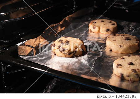 Chocolate chip cookies baked in the oven on foil. Focus and light on the individual cookie. The process of baking homemade biscuits. Close up. Minimalist professional photography. Horizontal image. 134423595