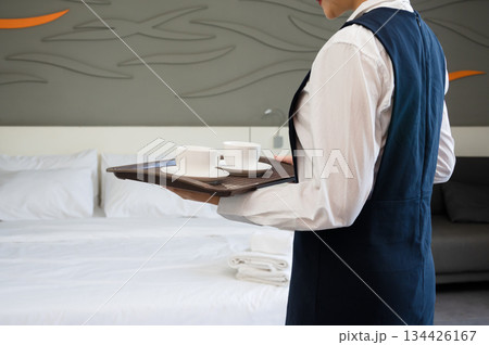 Hotel room service staff carrying tray with white coffee cups and saucers in modern clean suite with made bed Hotel room service staff carrying tray with white coffee cups and saucers in modern clean suite with made bed 134426167