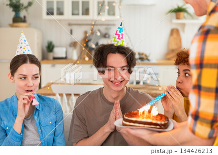 Make a wish. Man wearing party cap blowing out burning candles on birthday cake. Happy Birthday party. Group of friends wishes guy happy birthday. People celebrating birthday with party at home Make a wish. Man wearing party cap blowing out burning candles on birthday cake. Happy Birthday party. Group of friends wishes guy happy birthday. People celebrating birthday with party at home 134426213