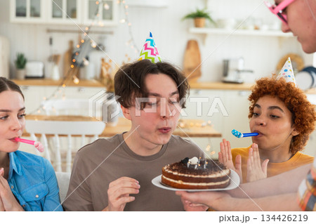Make a wish. Man wearing party cap blowing out burning candles on birthday cake. Happy Birthday party. Group of friends wishes guy happy birthday. People celebrating birthday with party at home Make a wish. Man wearing party cap blowing out burning candles on birthday cake. Happy Birthday party. Group of friends wishes guy happy birthday. People celebrating birthday with party at home 134426219