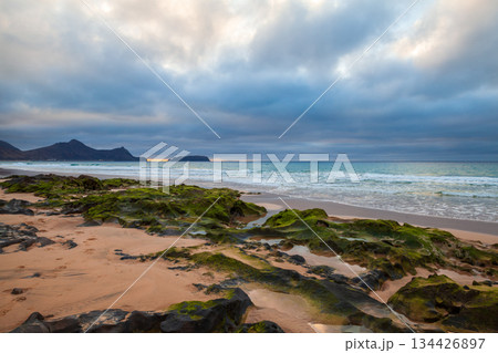 A serene beach scene with dark rocks, soft waves, and a moody, cloud filled sky 134426897