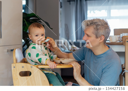 Young father feeding with a spoon adorable messy toddler baby sitting in high chair. Cute baby learning to eat food by himself. Kid enjoying healthy nutrition. family bonding moment during mealtime. Young father feeding with a spoon adorable messy toddler baby sitting in high chair. Cute baby learning to eat food by himself. Kid enjoying healthy nutrition. family bonding moment during mealtime. 134427158