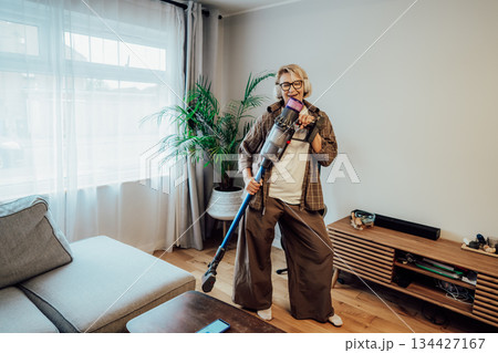 Middle aged woman singing and dancing with uplifting music in headphones while getting floor clean with cordless vacuum. Housewife having fun while doing household chores. Housekeeping, home cleaning Middle aged woman singing and dancing with uplifting music in headphones while getting floor clean with cordless vacuum. Housewife having fun while doing household chores. Housekeeping, home cleaning 134427167