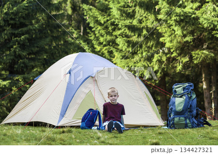 Holiday camping.Happy young boy sitting in front of a tent near backpacks taking rest after hiking in the forest.Traveling and tourism concept. Holiday camping.Happy young boy sitting in front of a tent near backpacks taking rest after hiking in the forest.Traveling and tourism concept. 134427321