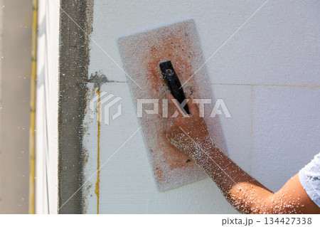 Hand of a worker with a tool for sanding and making smooth and even the surface of a house wall insulated with styrofoam insulation sheets. Hand of a worker with a tool for sanding and making smooth and even the surface of a house wall insulated with styrofoam insulation sheets. 134427338