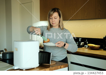 Woman uses tongs to serve food from white air fryer onto plate of rice. Smart air fryer, connected kitchen appliances, modern cooking, healthy eating. Woman uses tongs to serve food from white air fryer onto plate of rice. Smart air fryer, connected kitchen appliances, modern cooking, healthy eating. 134427453