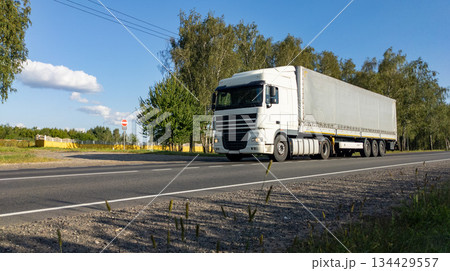 White semi-truck with a fully loaded cargo trailer driving along countryside road. The lorry travels on an empty freeway. White semi-truck with a fully loaded cargo trailer driving along countryside road. The lorry travels on an empty freeway. 134429557