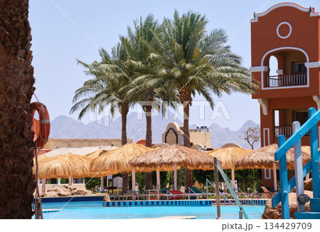 Empty deck chairs under straw shade umbrellas on swimming pool side in tropical resort. Summer vacations and getaway concept Empty deck chairs under straw shade umbrellas on swimming pool side in tropical resort. Summer vacations and getaway concept 134429709