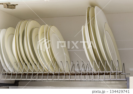 Dish drying metal rack with big nice white and light yellow clean plates on blurred background. Traditional comfortable kitchen. 134429741