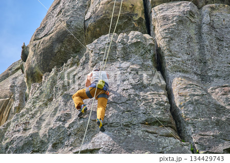Determined climber clambering up steep wall of rocky mountain. Sportsman overcoming difficult route. Engaging in extreme sports and rock climbing hobby concept. Determined climber clambering up steep wall of rocky mountain. Sportsman overcoming difficult route. Engaging in extreme sports and rock climbing hobby concept. 134429743