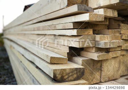 Close-up of piled stack of natural brown uneven rough wooden boards lit by bright sun. Industrial timber for carpentry, building, repairing and furniture, lumber material for construction. Close-up of piled stack of natural brown uneven rough wooden boards lit by bright sun. Industrial timber for carpentry, building, repairing and furniture, lumber material for construction. 134429898