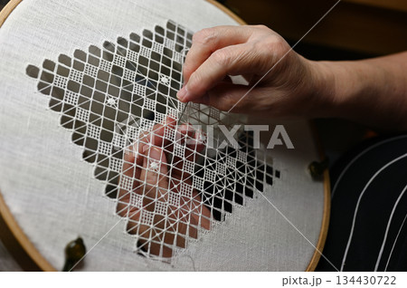 hands carefully stitching a delicate geometric lace pattern on white linen stretched in an embroidery hoop 134430722