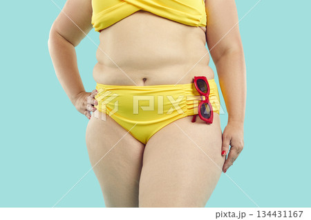 Crop shot of overweight woman with fat belly in yellow bikini standing on blue background 134431167
