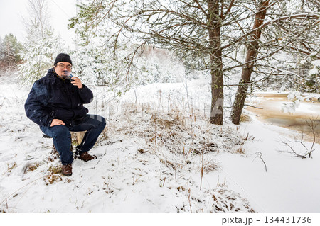 First snow has fallen, man in his 40s is smoking in forest by river, sitting on makeshift bench. 134431736