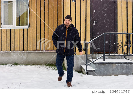 Emerging from wooden building with vertical siding, warmly dressed man over fifty moves into snowy yard amid falling snow during winter daylight. 134431747