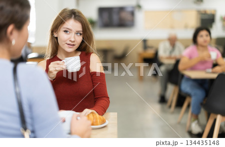 Young girl chatting with friend over coffee and croissant in cafeteria Young girl chatting with friend over coffee and croissant in cafeteria 134435148