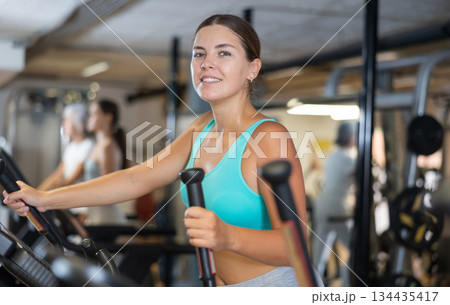 Slender athletic girl during cardio exercises on elliptical simulator in fitness center. 134435417